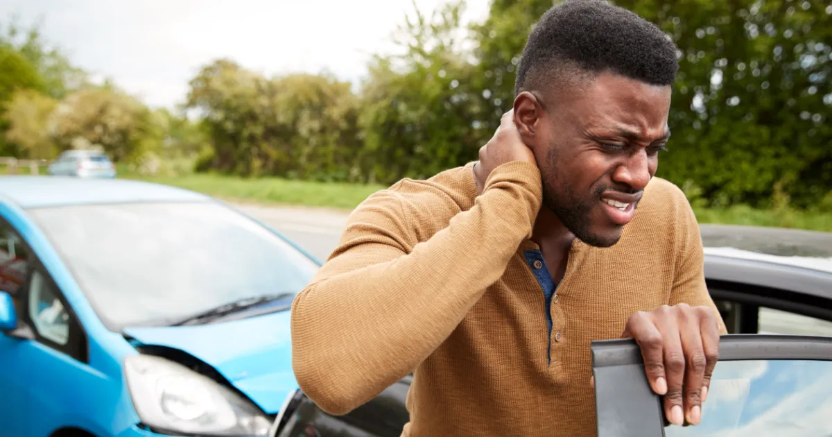 A man holding his neck in pain while standing next to a car involved in a traffic accident.
