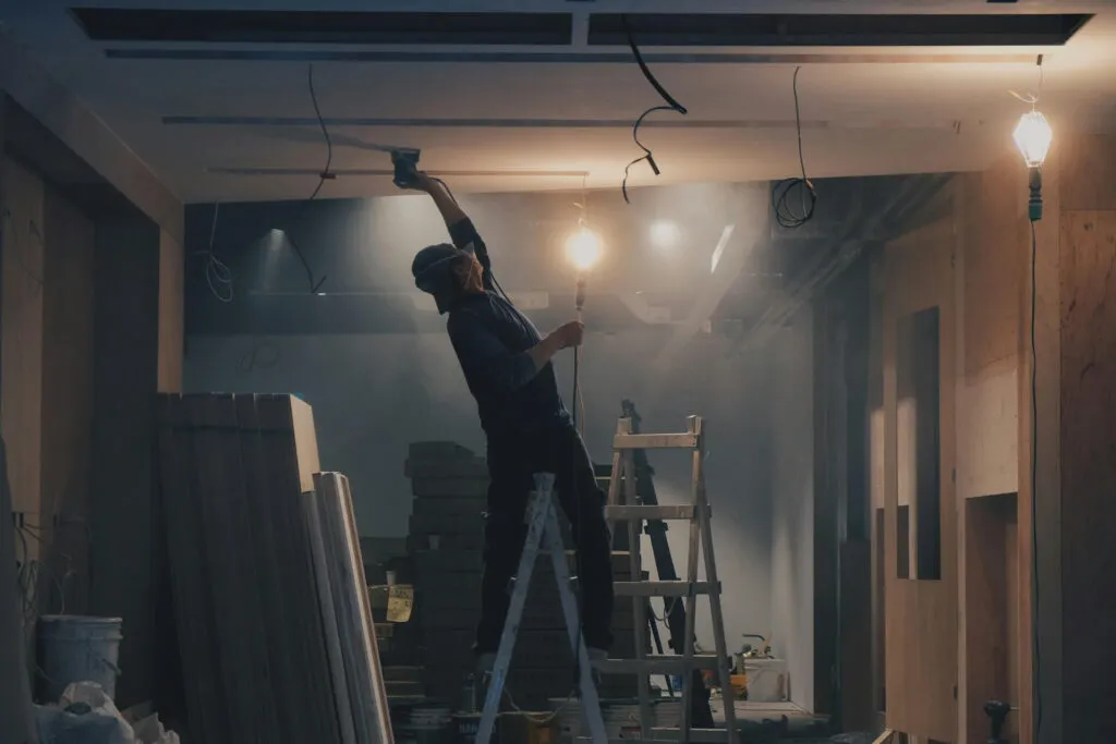 A person is standing on a step ladder, working on the ceiling of what appears to be a room undergoing construction or renovation.