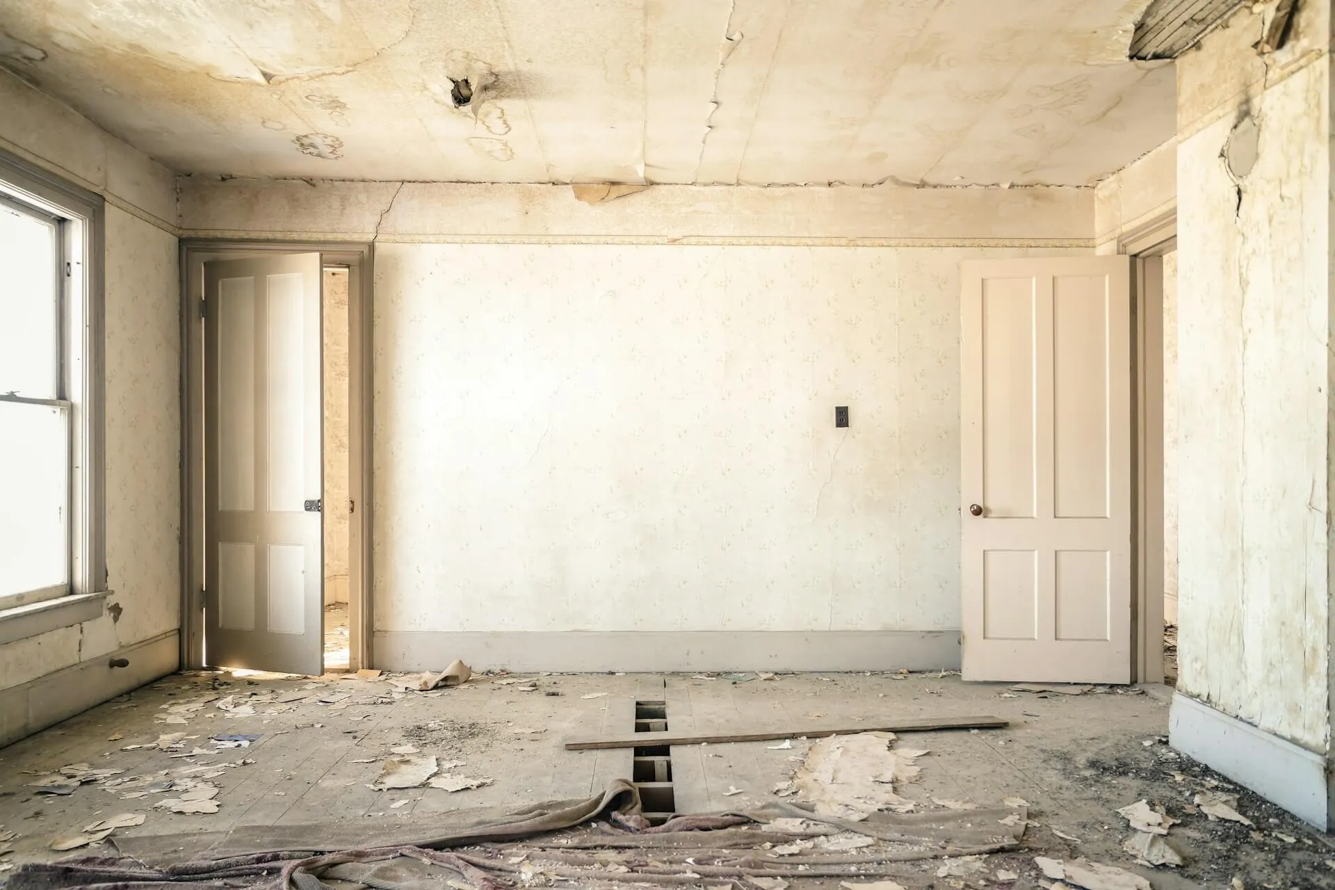An interior view of an empty, fixer-upper room featuring white walls, doorways, and a floor covered in debris and construction materials.