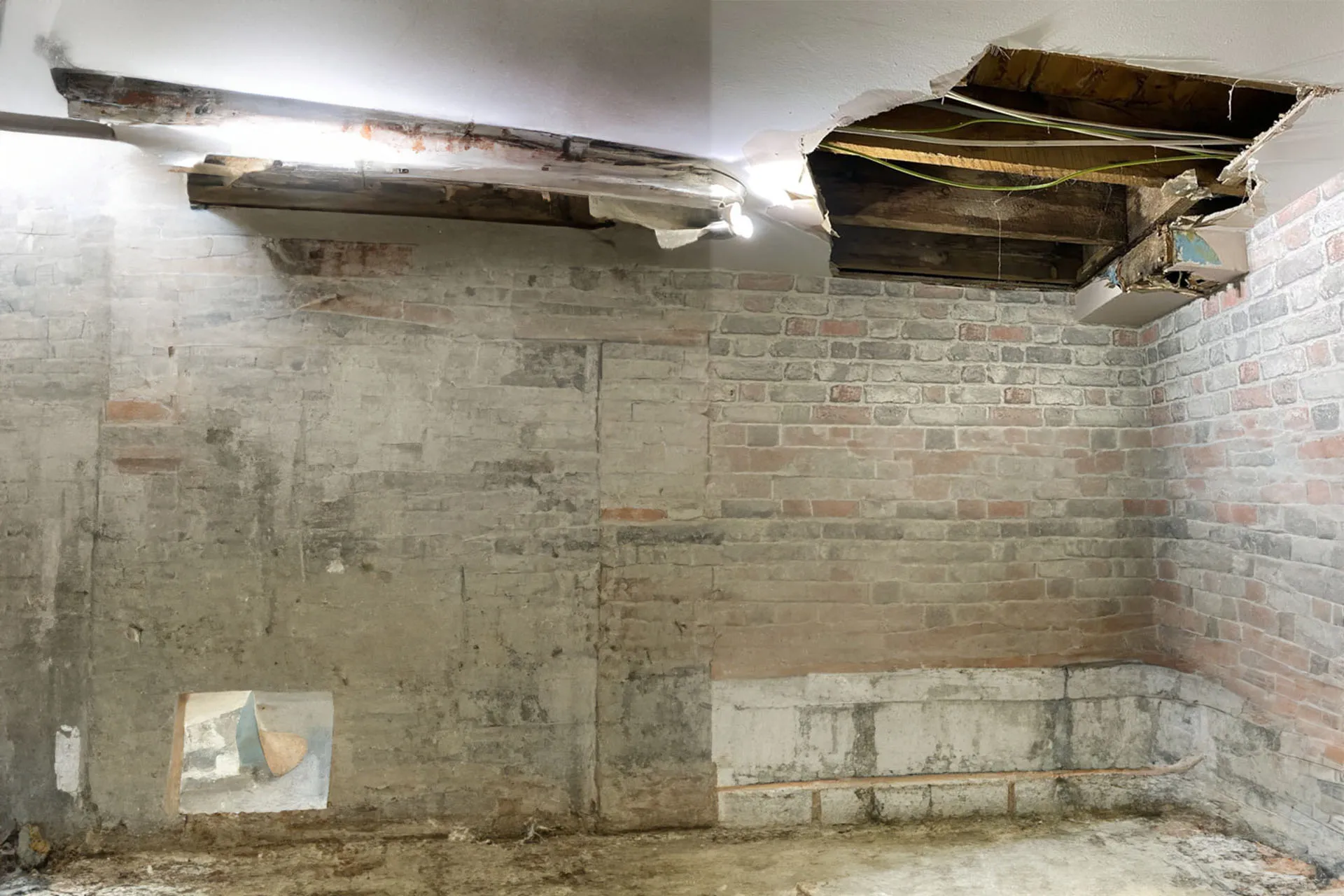 An interior view of a room undergoing demolition or heavy repairs, showing exposed concrete walls, raw masonry, and sections of the ceiling that have been torn down.