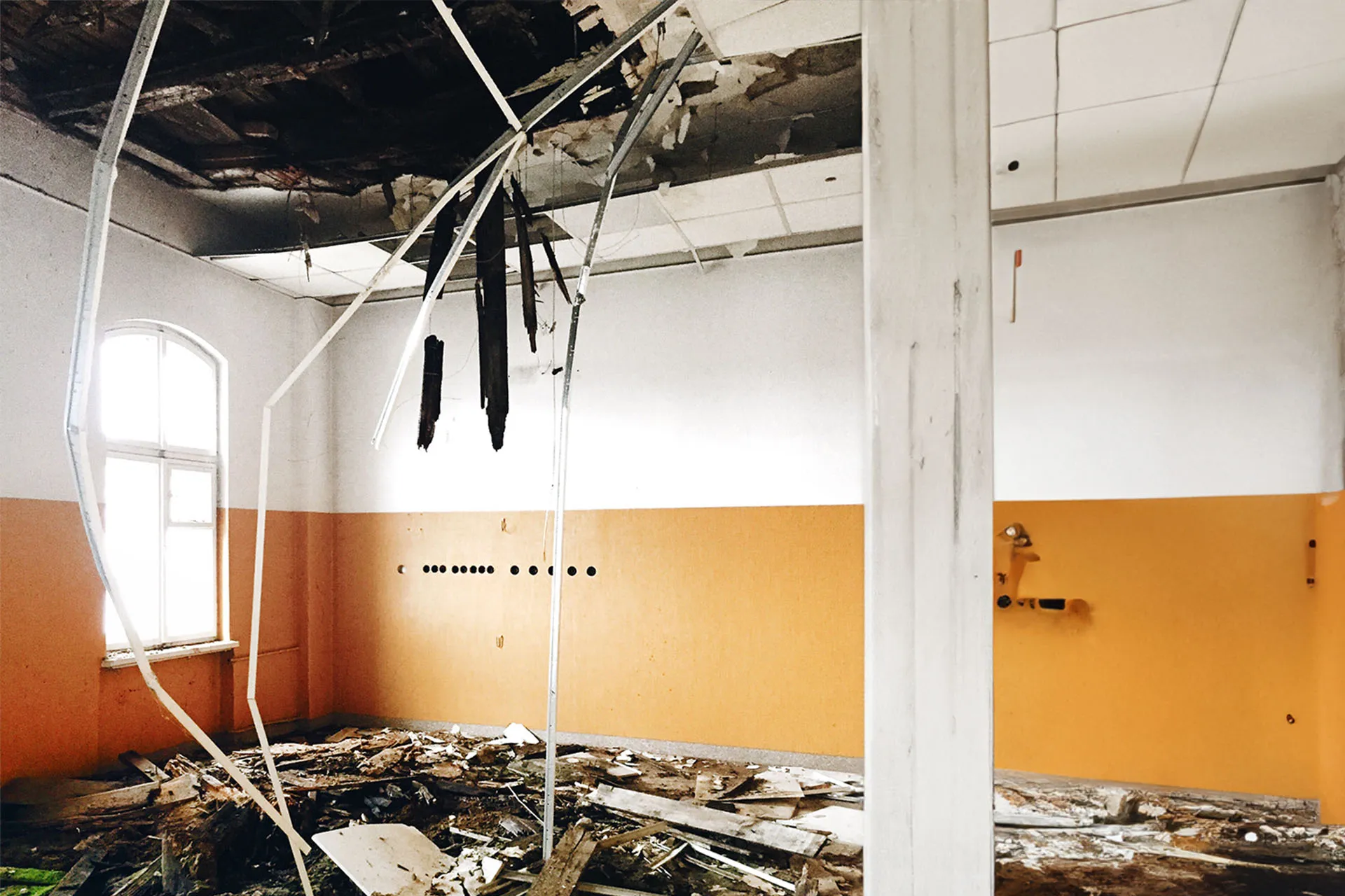An interior view of a damaged building room featuring a partially collapsed ceiling, debris scattered on the floor, and orange-painted walls.