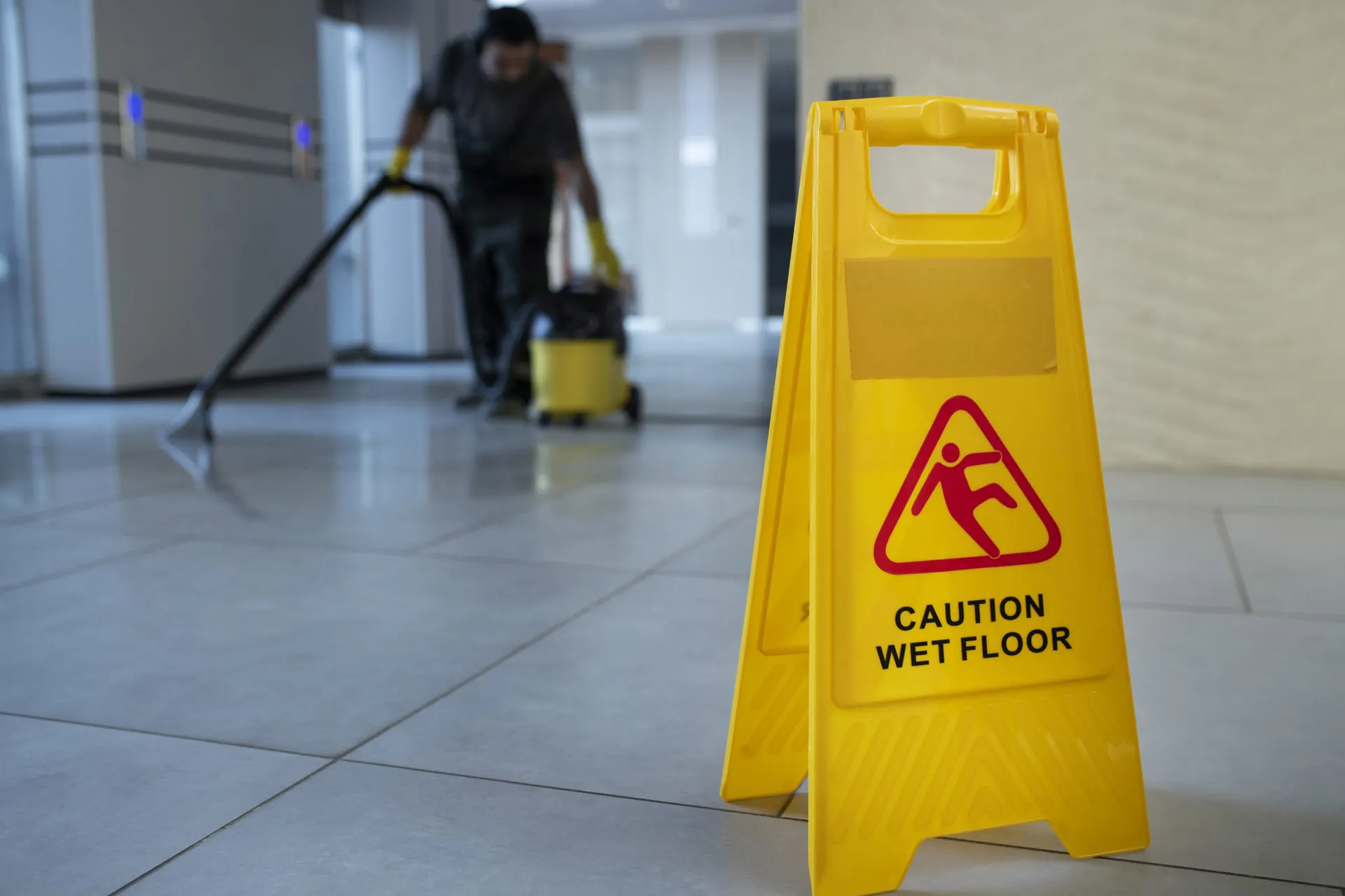 A yellow "Caution: Wet Floor" sign stands in the foreground while a person in the background cleans the floor with a vacuum.