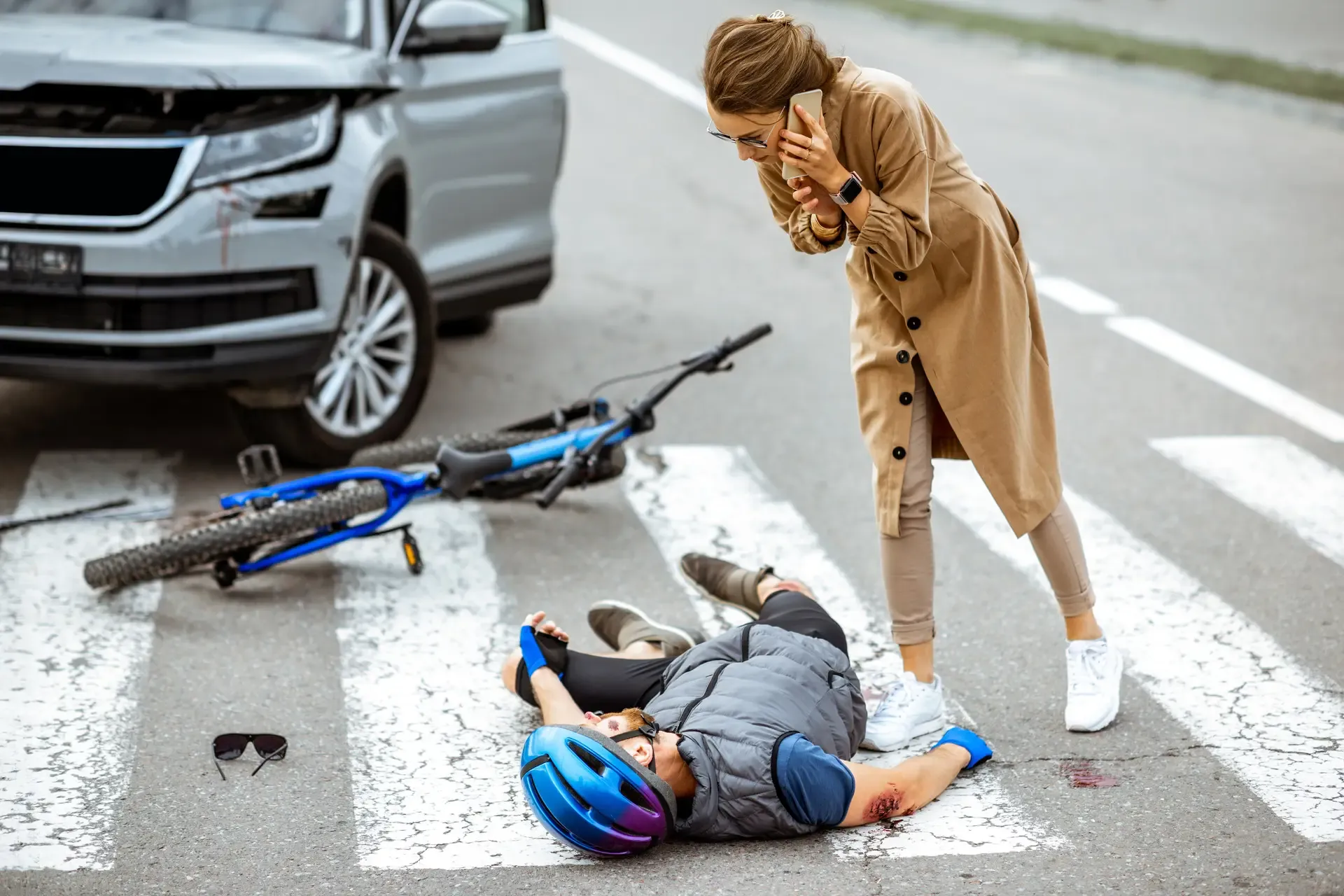 A distressed woman calling for help next to a cyclist lying on a crosswalk after a collision with a silver SUV.