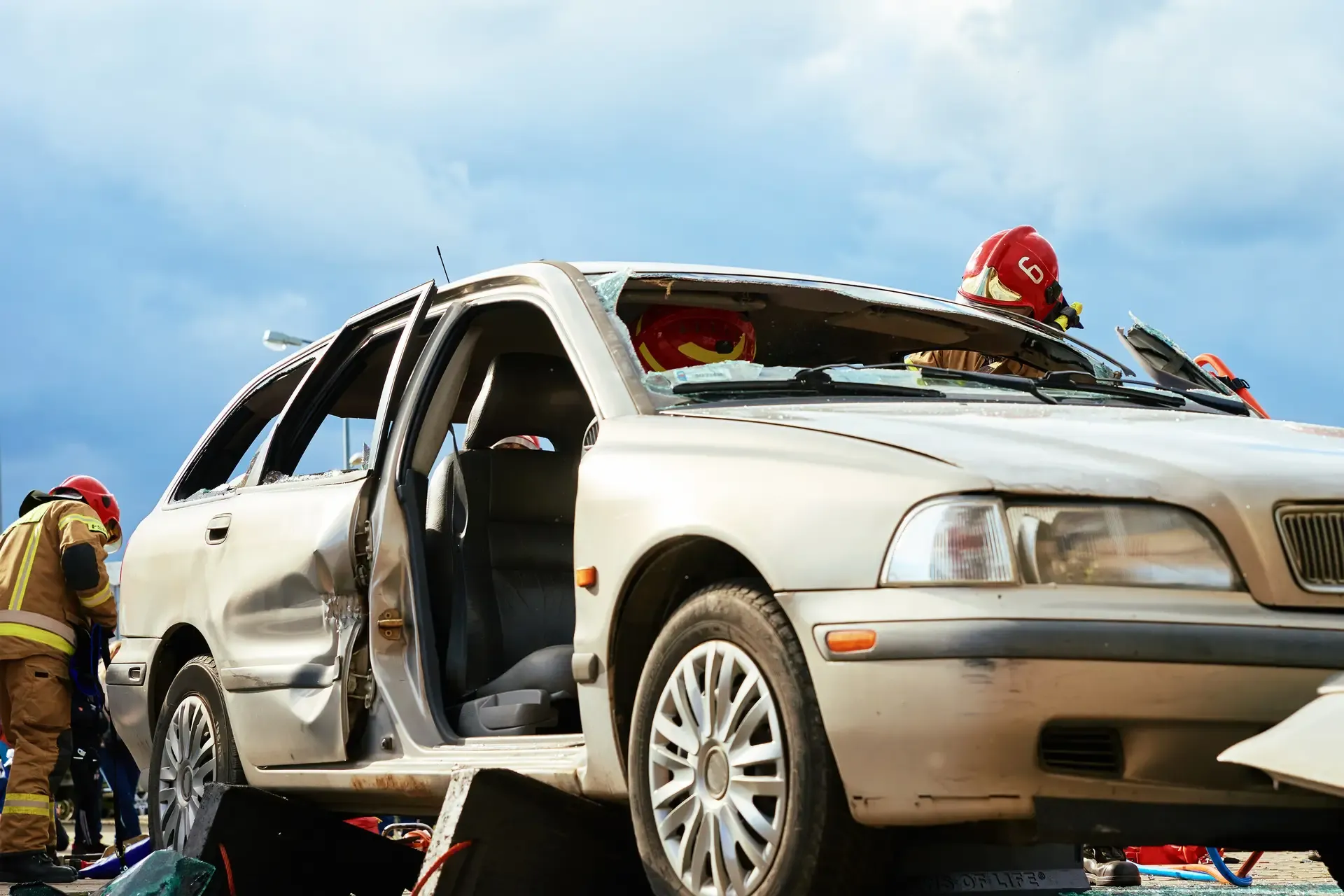 Emergency responders in helmets attending to a severely damaged silver car at an accident scene.