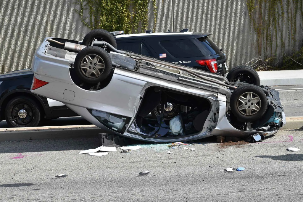 A silver car is overturned on its roof in the middle of a roadway, with police vehicles visible in the background.