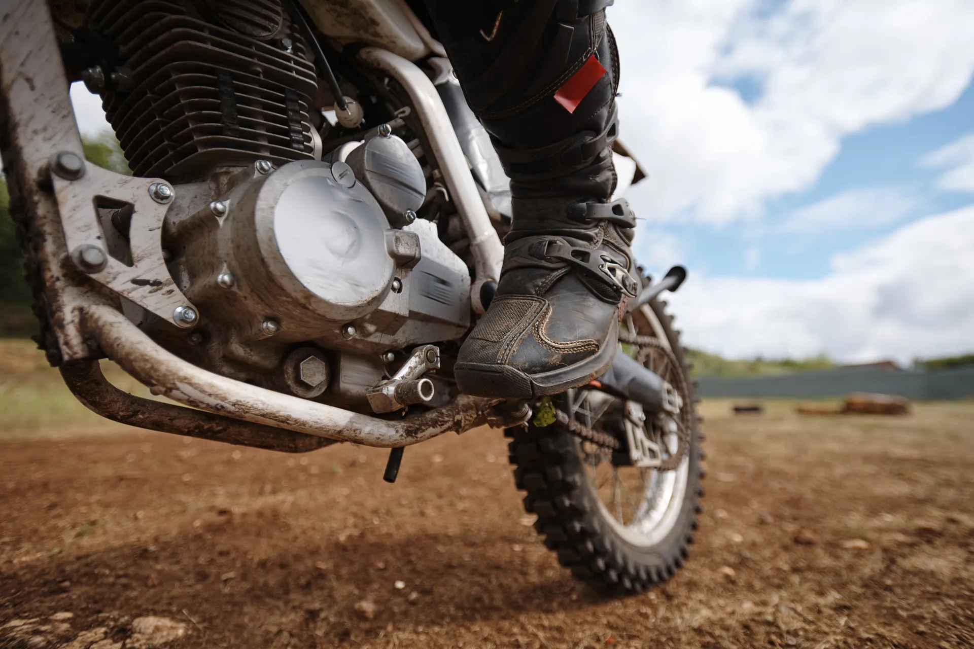 A close-up, low-angle view of a dirt bike's engine, frame, and rider's boot on a dirt track.
