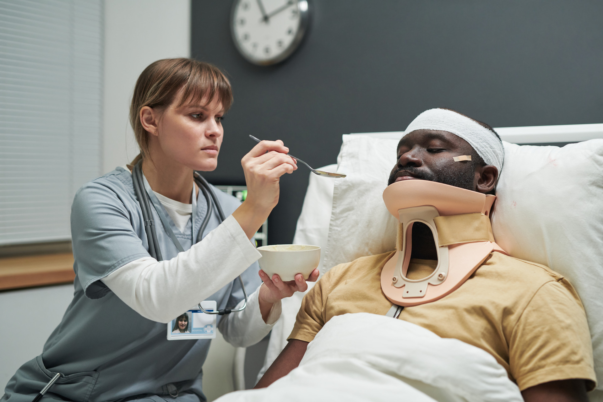 A healthcare professional in scrubs feeding a patient who is wearing a neck brace and a head bandage while lying in a hospital bed.
