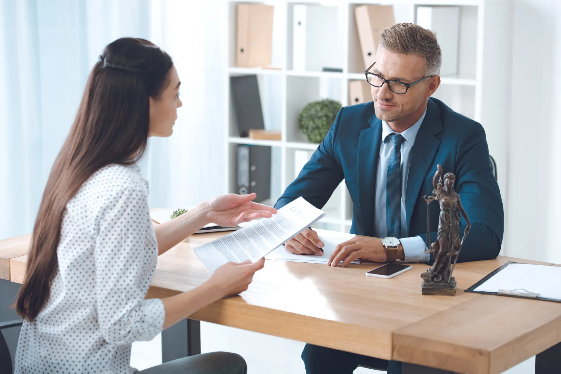 A male attorney in a suit and glasses is sitting at a wooden desk in his office, reviewing documents with a female client who is holding a paper and gesturing as they discuss a legal matter.