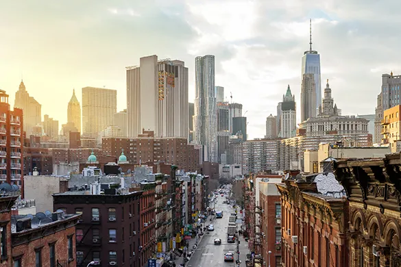 An urban cityscape during golden hour, featuring a street flanked by brick buildings leading toward a dense skyline of skyscrapers.