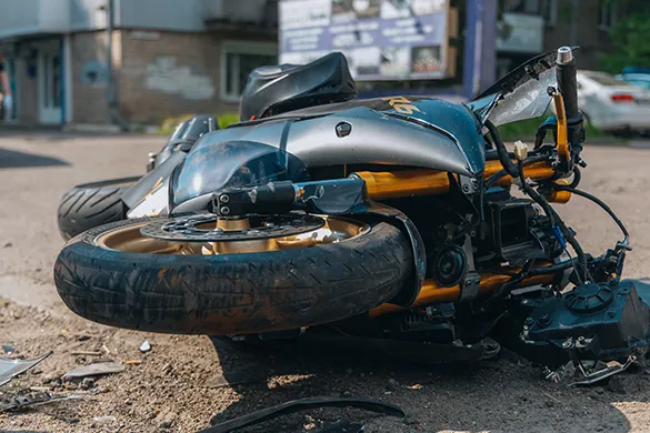 A damaged motorcycle lying on its side on a city street after an accident, with visible debris and residential buildings in the background.