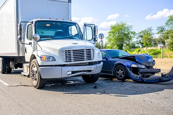 A white semi-truck and a blue sedan involved in a collision on a road, showing front-end damage to the car.
