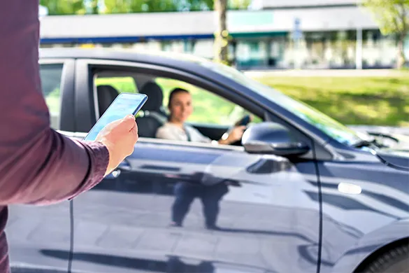 A person holding a smartphone in the foreground, with a woman sitting in the driver's seat of a parked car in the background, suggesting a ride-sharing or digital interaction.