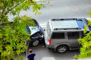 An overhead view of a car accident between a dark-colored sedan and a silver SUV on an asphalt road, partially framed by green foliage, with a person standing nearby.