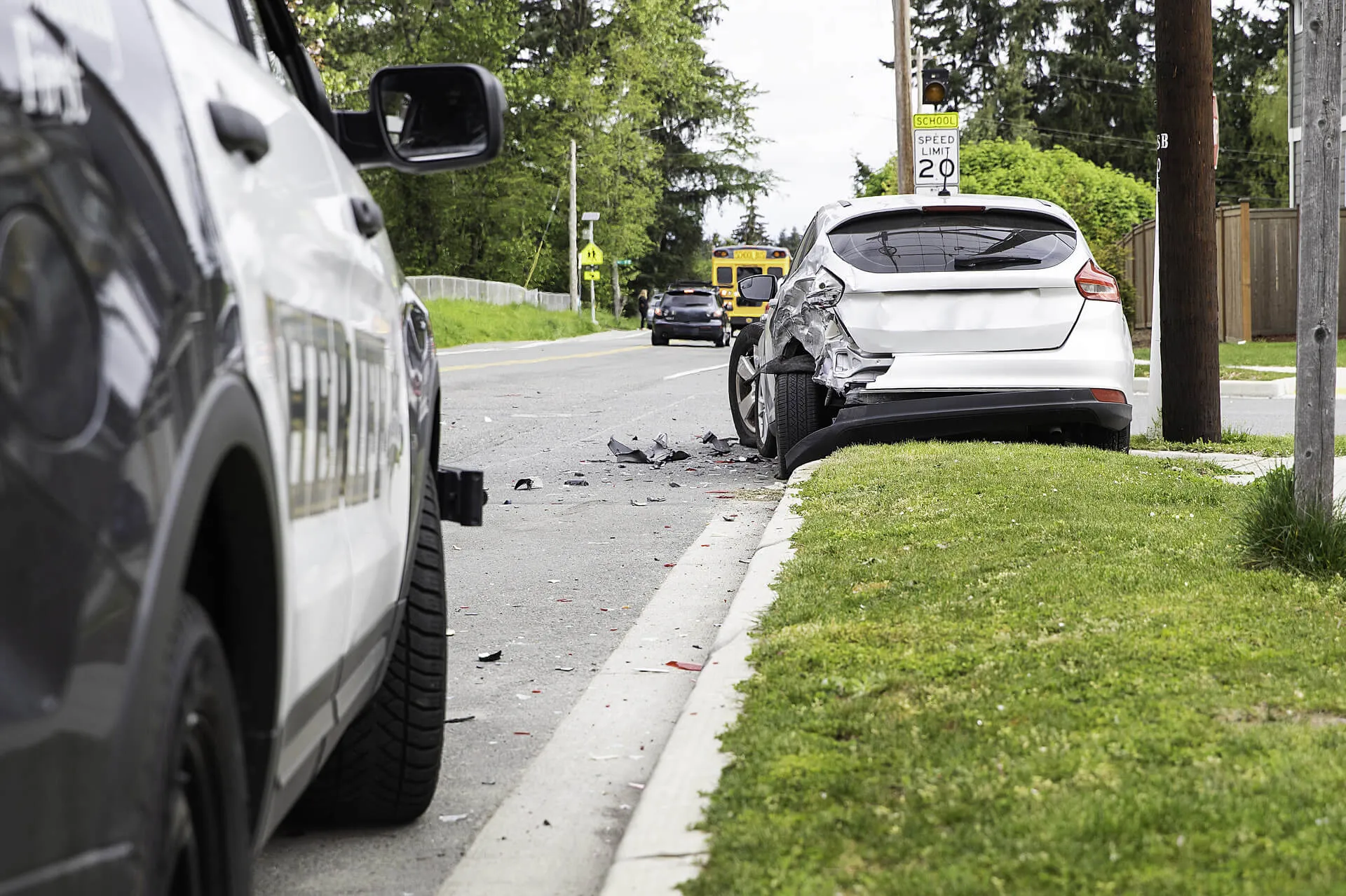 A white hatchback with significant rear-end damage sits on the shoulder of a suburban road following a car accident. Debris is scattered on the pavement behind the vehicle, and a law enforcement or emergency vehicle is visible in the foreground on the left, observing the scene.