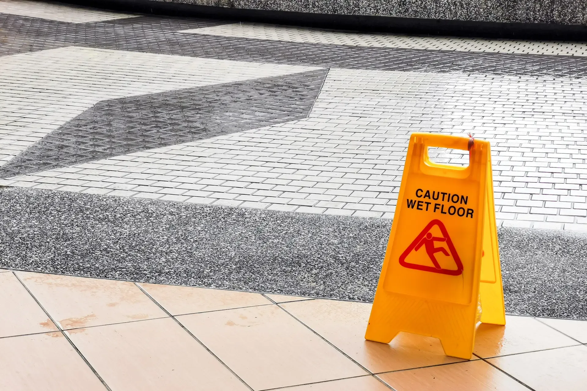 A bright yellow "Caution: Wet Floor" sign stands on a tiled surface, warning pedestrians of a potentially slippery area nearby. The sign features a standard black text warning and a red triangular icon depicting a person slipping.