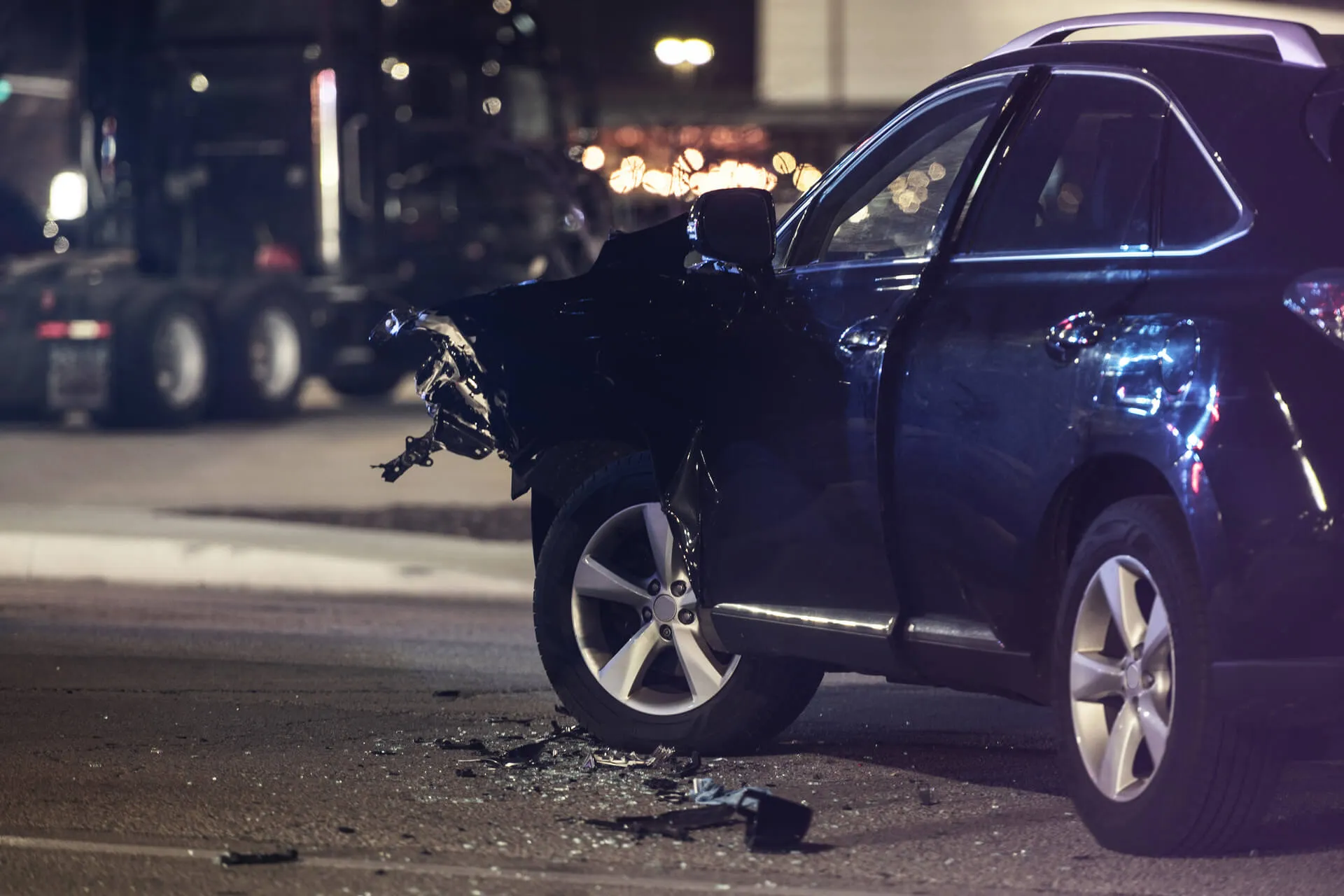 A dark-colored SUV is stopped on an asphalt road at night with severe damage to its front driver-side fender and bumper. Debris from the damaged area is scattered on the ground near the front wheel, and the vehicle appears to have been involved in a collision.